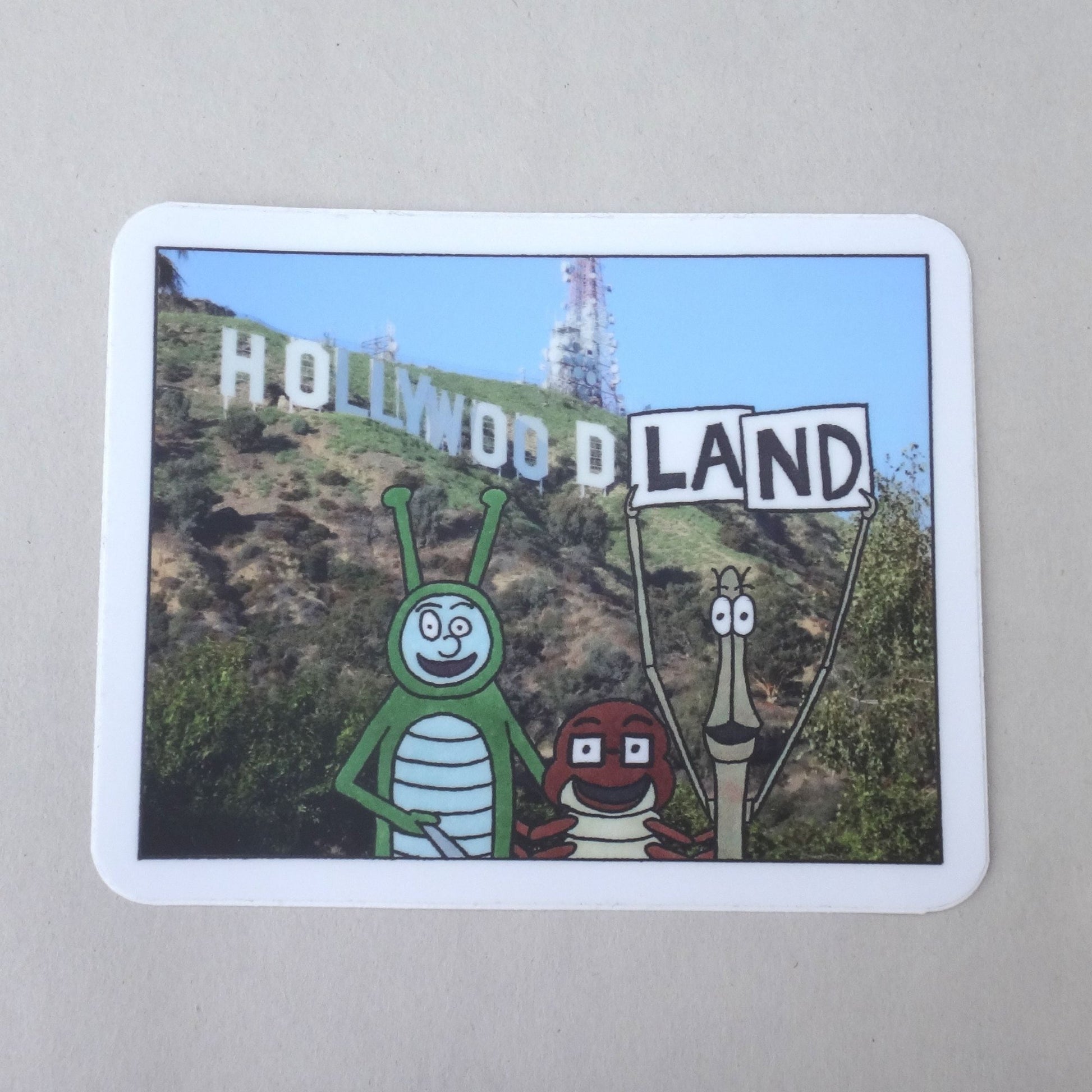 A sticker of Bug Boy, Seymour, and Will in front of the Hollywood sign. Will is holding up their own signs so it reads "Hollywoodland"