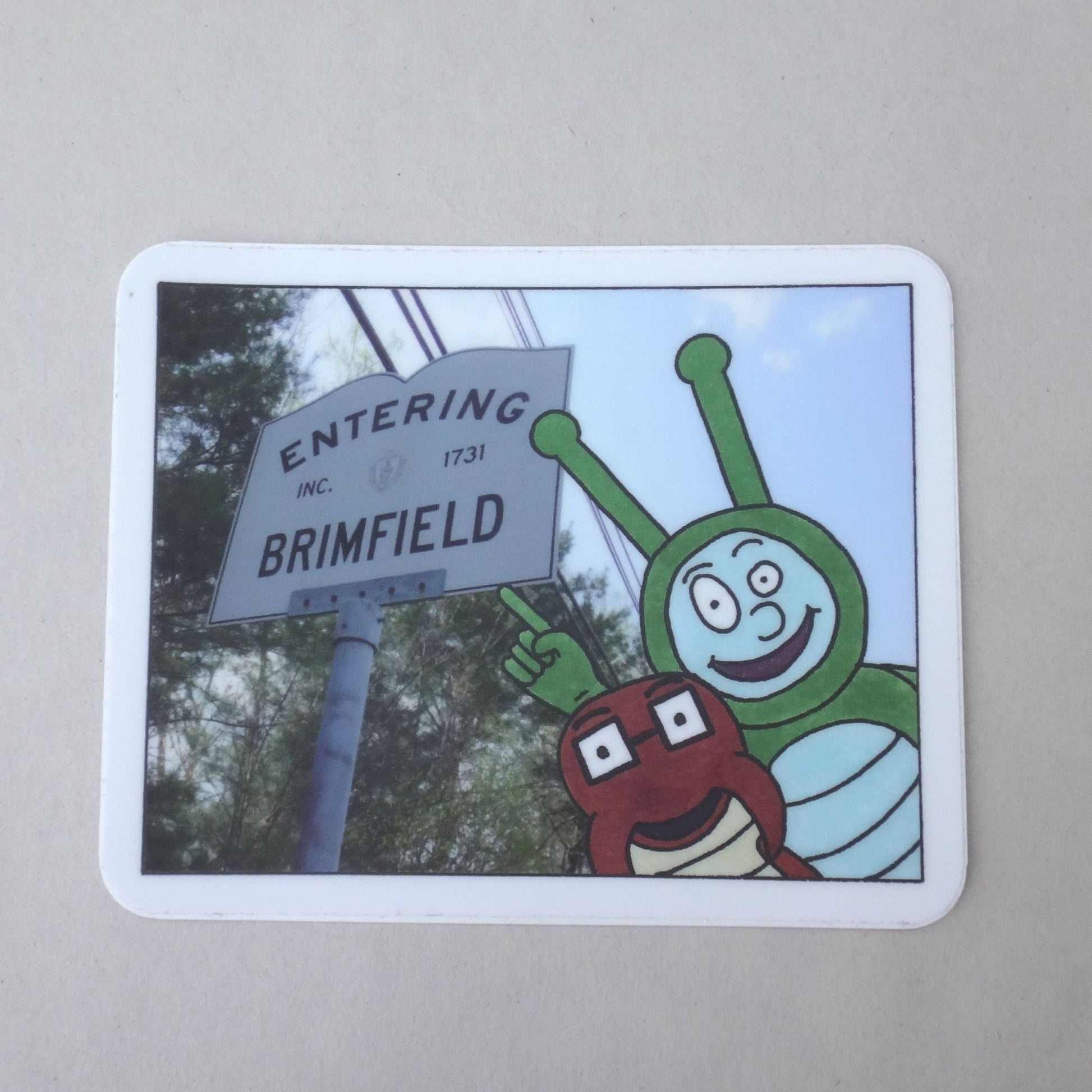 A sticker of Bug Boy & Seymour in front of the Brimfield, MA sign